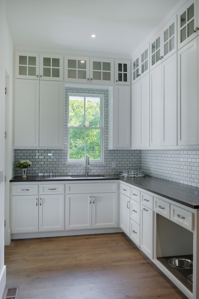 This is an example of a mid-sized country u-shaped kitchen pantry in Minneapolis with a farmhouse sink, shaker cabinets, white cabinets, white splashback, subway tile splashback, stainless steel appliances, light hardwood floors, with island and brown floor.