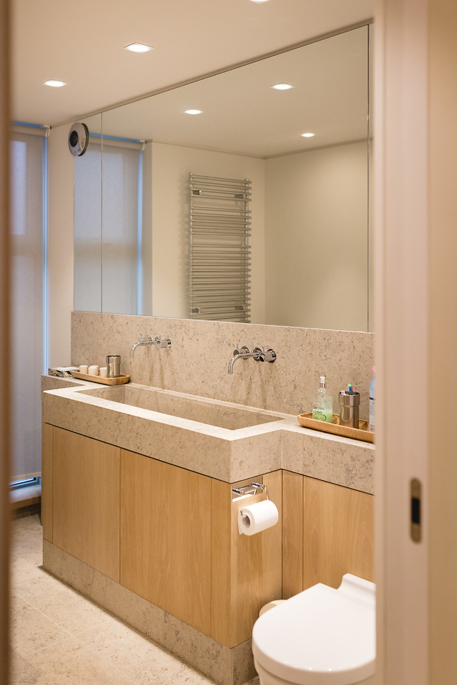 Mid-sized contemporary kids bathroom in London with flat-panel cabinets, light wood cabinets, a drop-in tub, a shower/bathtub combo, a wall-mount toilet, beige tile, stone slab, white walls, limestone floors, a trough sink, limestone benchtops, beige floor, a hinged shower door and beige benchtops.
