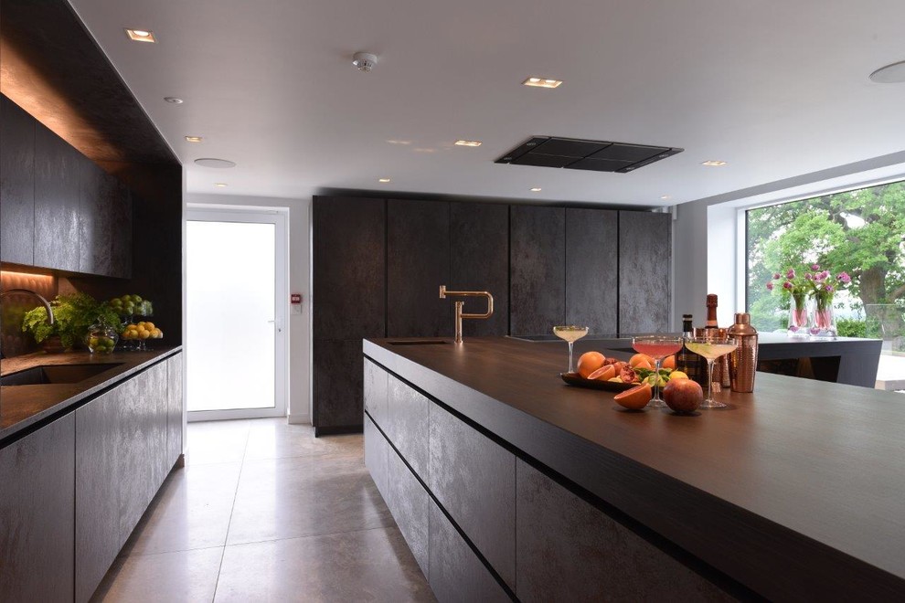 Expansive contemporary l-shaped kitchen in Manchester with a drop-in sink, flat-panel cabinets, distressed cabinets, solid surface benchtops, metallic splashback, metal splashback, black appliances, ceramic floors, with island and grey floor.