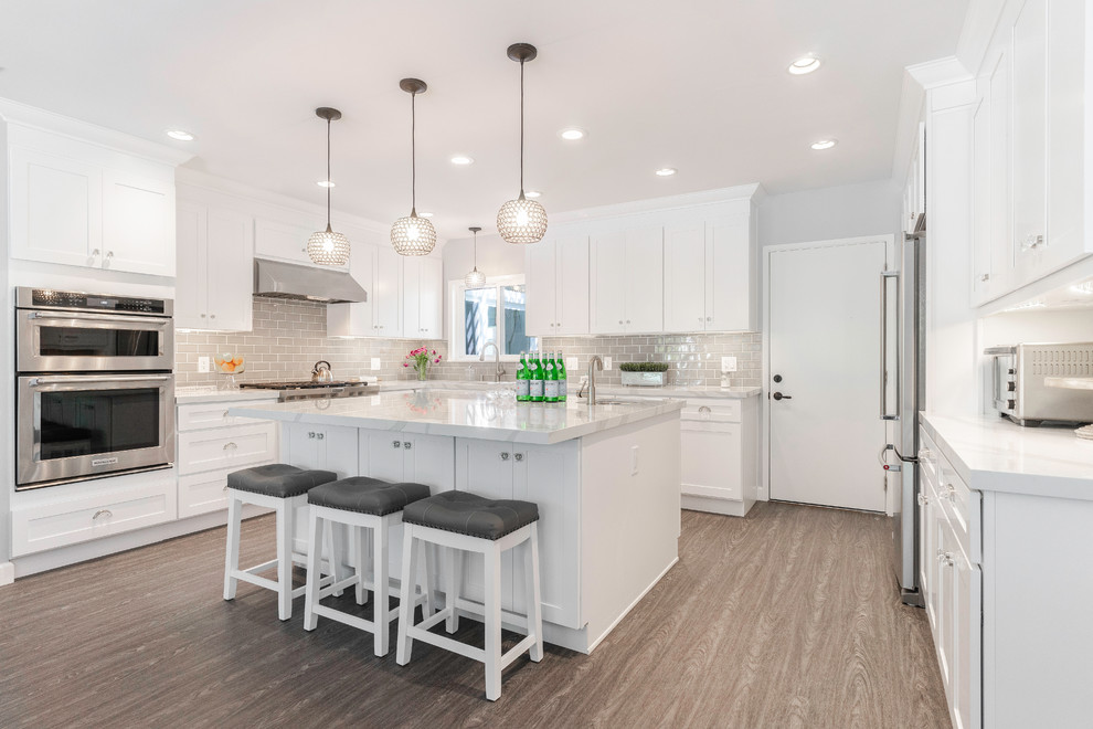 This is an example of a transitional u-shaped kitchen in Los Angeles with shaker cabinets, white cabinets, grey splashback, subway tile splashback, stainless steel appliances, with island, brown floor and white benchtop.