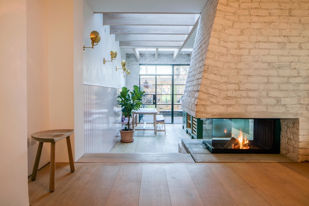 This is an example of a scandinavian open concept living room in London with pink walls, medium hardwood floors, a two-sided fireplace, a brick fireplace surround and no tv.