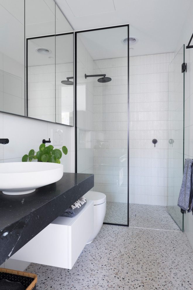 Photo of a mid-sized industrial bathroom in Melbourne with glass-front cabinets, white cabinets, a corner shower, a wall-mount toilet, white tile, ceramic tile, white walls, terrazzo floors, a vessel sink, marble benchtops, multi-coloured floor, an open shower, black benchtops, a single vanity and a built-in vanity.