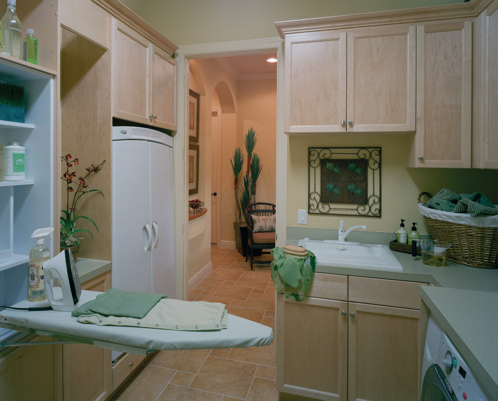 This is an example of a large mediterranean galley dedicated laundry room in Miami with a drop-in sink, recessed-panel cabinets, light wood cabinets, laminate benchtops, travertine floors, a side-by-side washer and dryer and beige walls.
