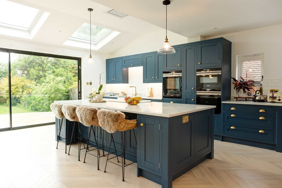 Photo of a transitional galley kitchen in Surrey with shaker cabinets, blue cabinets, white splashback, black appliances, light hardwood floors, with island, beige floor and white benchtop.