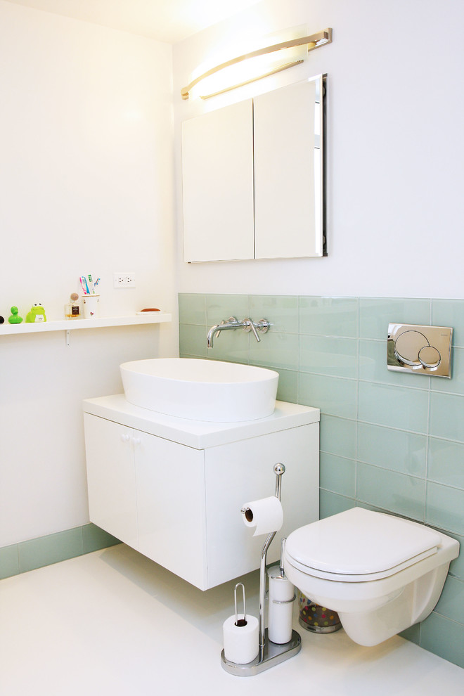 This is an example of a contemporary bathroom in New York with a vessel sink, flat-panel cabinets, white cabinets, a wall-mount toilet, blue tile, glass tile and white walls.