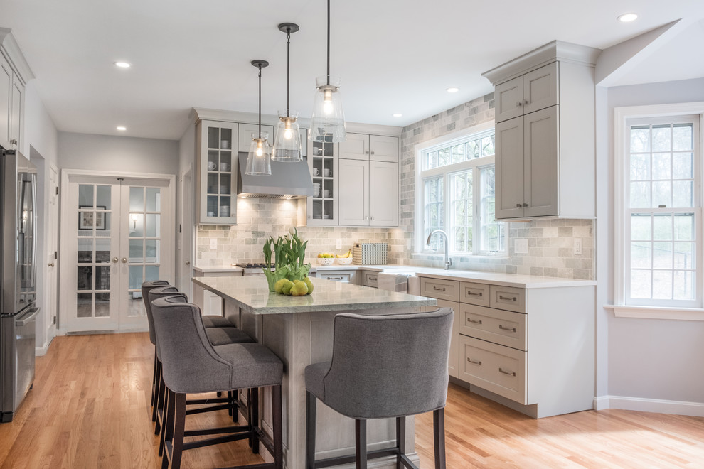 Mid-sized transitional l-shaped open plan kitchen in Boston with a farmhouse sink, recessed-panel cabinets, grey cabinets, quartz benchtops, green splashback, marble splashback, stainless steel appliances, light hardwood floors, with island and white benchtop.