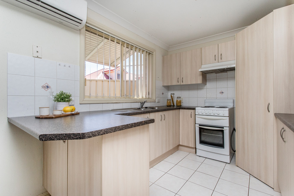 Small contemporary u-shaped open plan kitchen in Sydney with a double-bowl sink, flat-panel cabinets, beige cabinets, laminate benchtops, white splashback, ceramic splashback, white appliances, ceramic floors, a peninsula, white floor and brown benchtop.