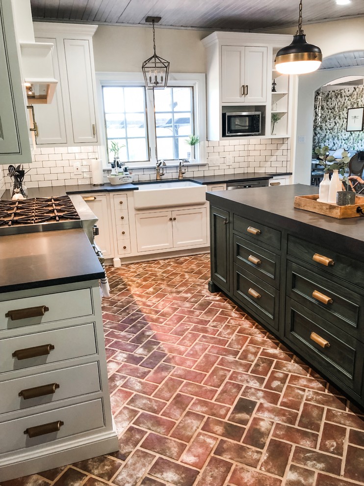 This is an example of a mid-sized country u-shaped eat-in kitchen in Philadelphia with a farmhouse sink, recessed-panel cabinets, white cabinets, soapstone benchtops, white splashback, subway tile splashback, stainless steel appliances, brick floors, with island, red floor and black benchtop.