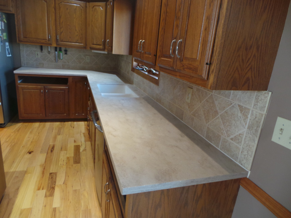 Large trendy l-shaped light wood floor and brown floor enclosed kitchen photo with an undermount sink, raised-panel cabinets, medium tone wood cabinets, solid surface countertops, beige backsplash, ceramic backsplash, stainless steel appliances, an island and beige countertops