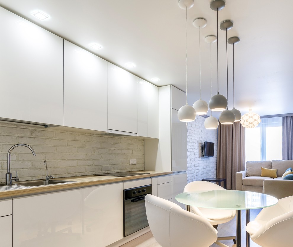 Inspiration for a mid-sized contemporary single-wall eat-in kitchen in Saint Petersburg with an undermount sink, flat-panel cabinets, white cabinets, wood benchtops, white splashback, brick splashback, stainless steel appliances, light hardwood floors and grey floor.