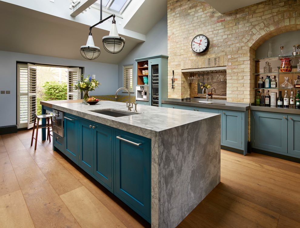 This is an example of a large transitional u-shaped separate kitchen in London with an undermount sink, recessed-panel cabinets, blue cabinets, grey splashback, stone slab splashback, stainless steel appliances, medium hardwood floors, with island, brown floor and quartzite benchtops.