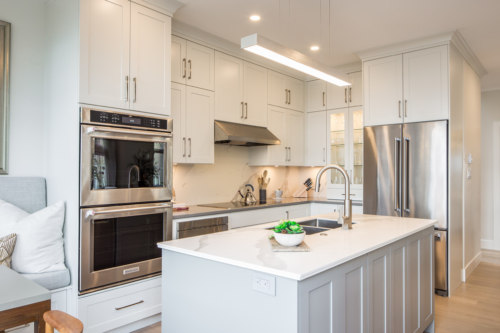 Inspiration for a transitional l-shaped kitchen in Vancouver with shaker cabinets, white cabinets, quartz benchtops, white splashback, porcelain splashback, stainless steel appliances, light hardwood floors, with island, beige floor, white benchtop and a double-bowl sink.