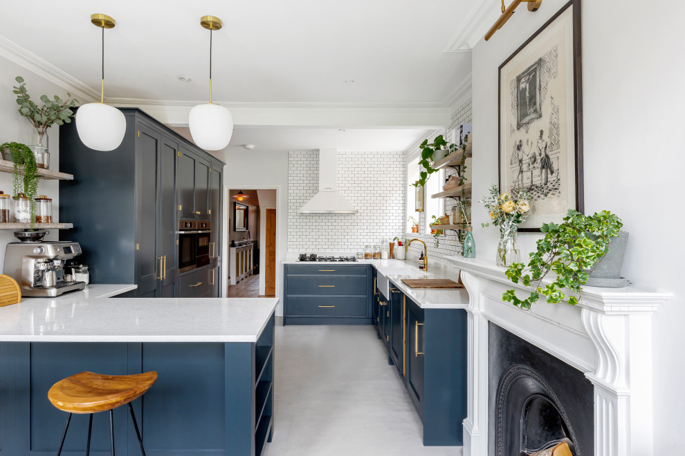 Photo of a large transitional l-shaped open plan kitchen in Other with a farmhouse sink, shaker cabinets, blue cabinets, solid surface benchtops, white splashback, subway tile splashback, concrete floors, a peninsula, grey floor, white benchtop and panelled appliances.