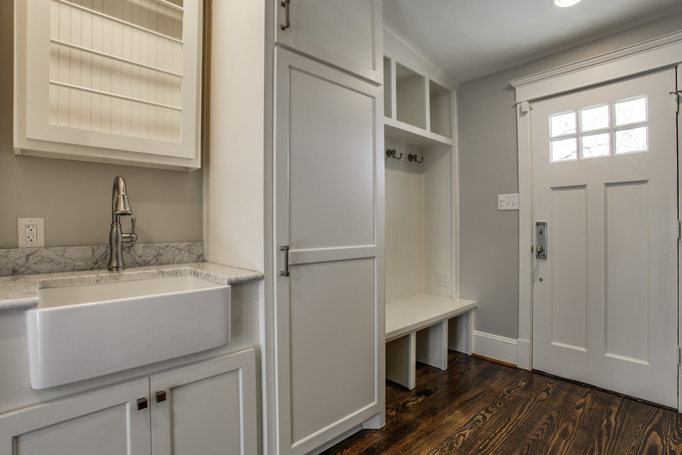 This is an example of a large transitional galley utility room in Dallas with a farmhouse sink, shaker cabinets, white cabinets, marble benchtops, grey walls, dark hardwood floors and a stacked washer and dryer.