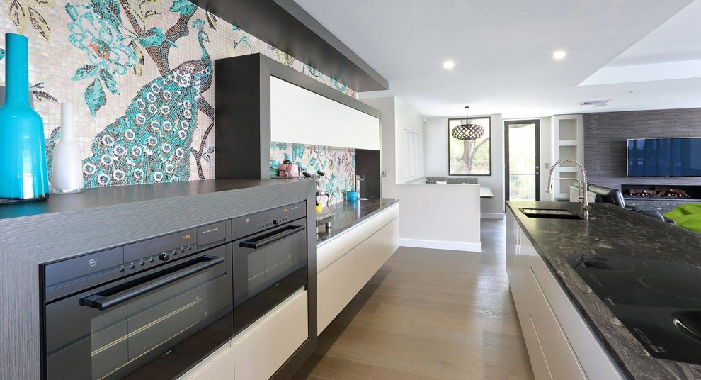 Design ideas for a mid-sized eclectic single-wall open plan kitchen in Brisbane with flat-panel cabinets, white cabinets, granite benchtops, multi-coloured splashback, mosaic tile splashback, stainless steel appliances, medium hardwood floors, with island and grey floor.