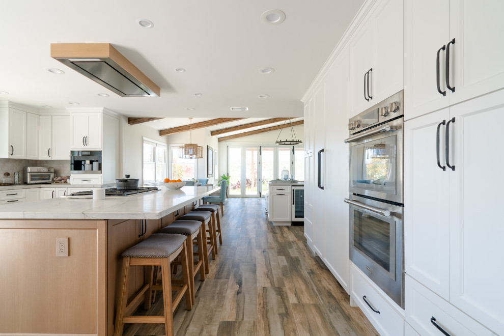 Inspiration for a large coastal galley porcelain tile, brown floor and exposed beam open concept kitchen remodel in Denver with a farmhouse sink, recessed-panel cabinets, white cabinets, quartzite countertops, white backsplash, ceramic backsplash, stainless steel appliances, an island and white countertops