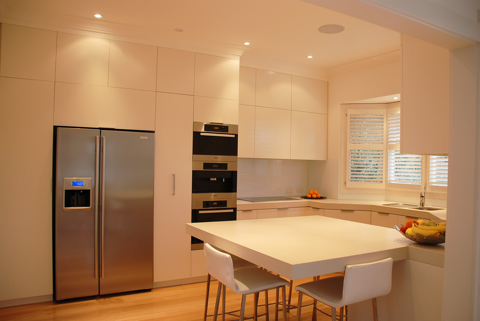 Mid-sized contemporary u-shaped eat-in kitchen in Sydney with a double-bowl sink, flat-panel cabinets, white cabinets, quartz benchtops, white splashback, glass sheet splashback, stainless steel appliances, light hardwood floors and a peninsula.