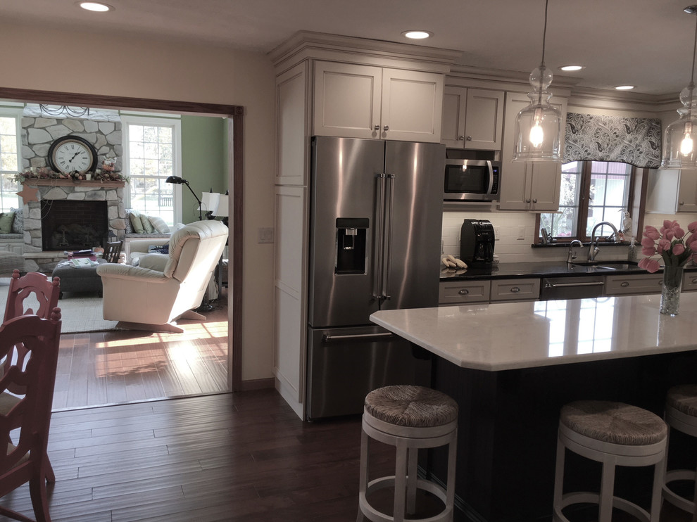 Example of a large transitional medium tone wood floor and brown floor eat-in kitchen design with an undermount sink, flat-panel cabinets, white cabinets, granite countertops, white backsplash, porcelain backsplash, stainless steel appliances and an island