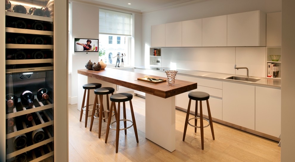 Photo of a mid-sized contemporary single-wall kitchen in London with an integrated sink, white splashback, medium hardwood floors, with island, flat-panel cabinets, white cabinets, stainless steel benchtops and glass sheet splashback.