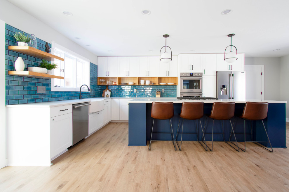 Expansive contemporary l-shaped open plan kitchen in Milwaukee with a farmhouse sink, shaker cabinets, white cabinets, quartz benchtops, blue splashback, porcelain splashback, stainless steel appliances, vinyl floors, white benchtop, with island and beige floor.