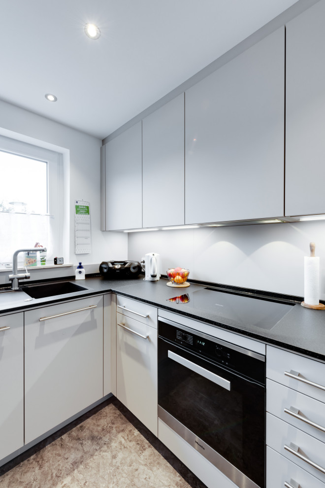 Mid-sized contemporary l-shaped separate kitchen in Hamburg with a single-bowl sink, flat-panel cabinets, white cabinets, laminate benchtops, white splashback, glass sheet splashback, black appliances, linoleum floors, a peninsula, grey floor, black benchtop and coffered.