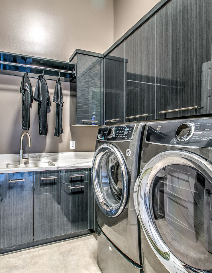 This is an example of a large transitional l-shaped dedicated laundry room in Omaha with a single-bowl sink, flat-panel cabinets, solid surface benchtops, beige walls, porcelain floors, a side-by-side washer and dryer and grey cabinets.