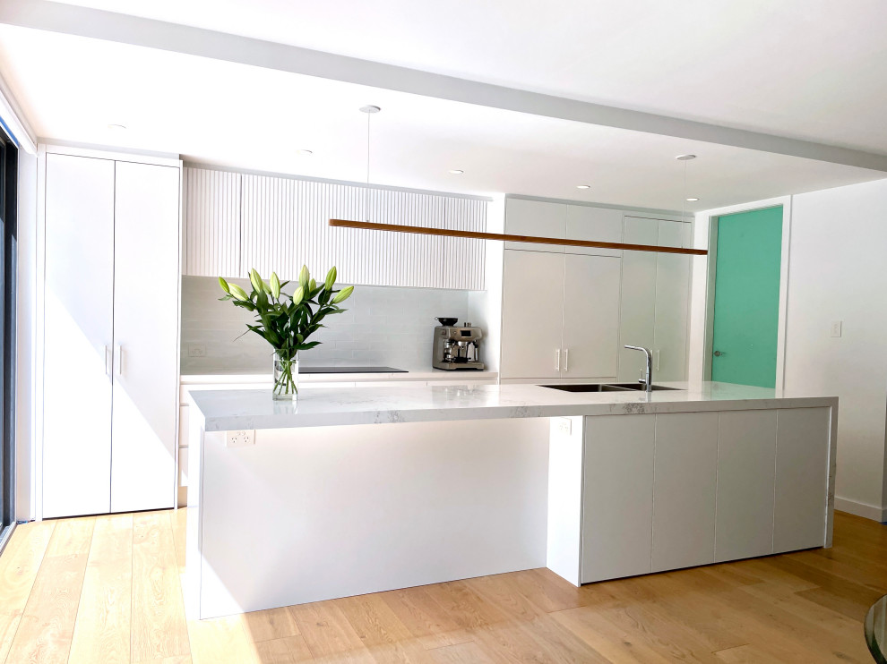 Design ideas for a large single-wall open plan kitchen in Sydney with an undermount sink, flat-panel cabinets, white cabinets, quartz benchtops, white splashback, subway tile splashback, black appliances, light hardwood floors, with island, brown floor and white benchtop.