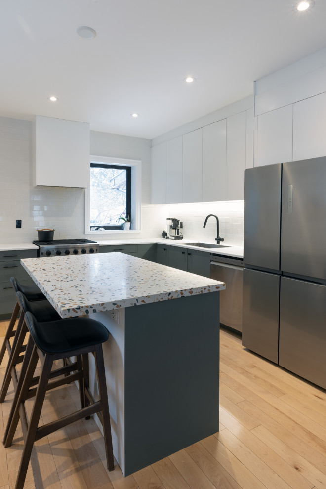 Inspiration for a medium sized contemporary l-shaped kitchen/diner in Toronto with a built-in sink, flat-panel cabinets, green cabinets, terrazzo worktops, white splashback, ceramic splashback, stainless steel appliances, light hardwood flooring and an island.