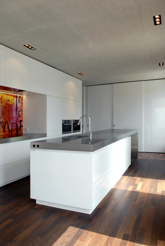 Photo of a large contemporary open plan kitchen in Dusseldorf with flat-panel cabinets, white cabinets, an integrated sink, stainless steel benchtops, multi-coloured splashback, stainless steel appliances, dark hardwood floors and with island.