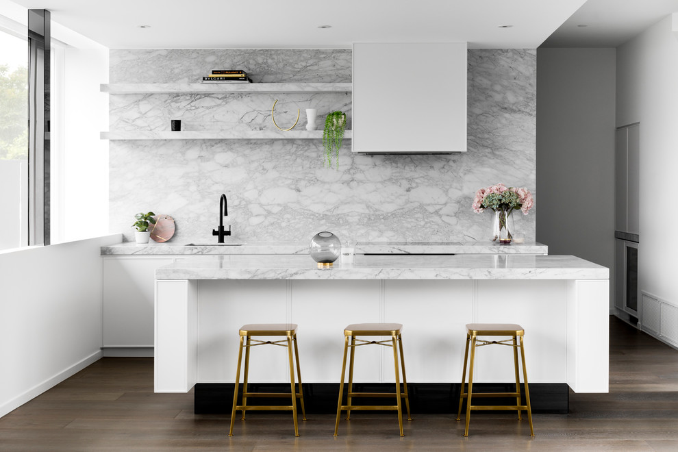 Photo of a contemporary galley kitchen in Melbourne with an undermount sink, flat-panel cabinets, white cabinets, marble benchtops, white splashback, marble splashback, black appliances, medium hardwood floors, with island, brown floor and white benchtop.