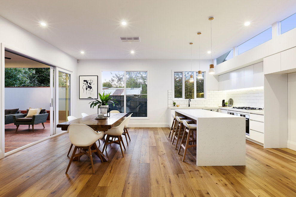 This is an example of an expansive modern l-shaped eat-in kitchen in Melbourne with an undermount sink, flat-panel cabinets, white cabinets, quartz benchtops, white splashback, subway tile splashback, stainless steel appliances, light hardwood floors, with island, brown floor and white benchtop.