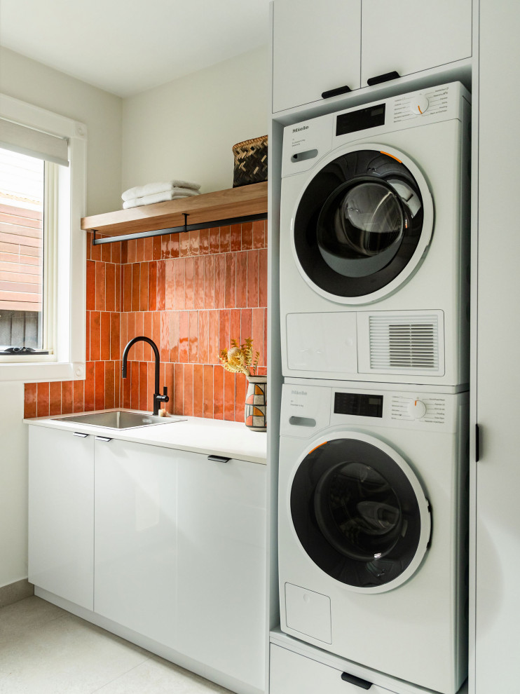 Photo of a mid-sized contemporary galley dedicated laundry room in Melbourne with a drop-in sink, flat-panel cabinets, white cabinets, quartz benchtops, orange splashback, ceramic splashback, white walls, porcelain floors, a stacked washer and dryer, beige floor and white benchtop.