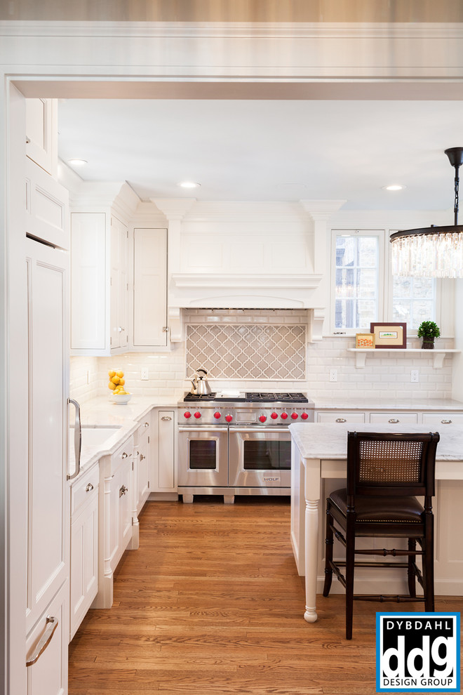 Klassisk inredning av ett avskilt, stort kök, med en nedsänkt diskho, luckor med profilerade fronter, vita skåp, marmorbänkskiva, vitt stänkskydd, stänkskydd i tunnelbanekakel, integrerade vitvaror, mellanmörkt trägolv, en köksö och brunt golv