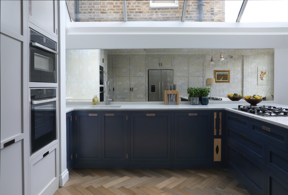 This is an example of a mid-sized transitional u-shaped eat-in kitchen in London with an undermount sink, shaker cabinets, blue cabinets, quartzite benchtops, mirror splashback, stainless steel appliances, medium hardwood floors and no island.