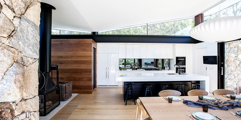 Photo of a mid-sized contemporary galley eat-in kitchen in Melbourne with an undermount sink, white cabinets, solid surface benchtops, mirror splashback, black appliances, medium hardwood floors and with island.