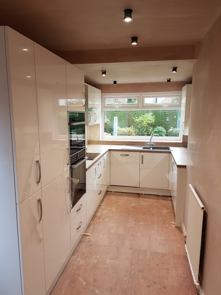 Small contemporary galley separate kitchen in Manchester with a drop-in sink, flat-panel cabinets, white cabinets, wood benchtops, blue splashback, panelled appliances and vinyl floors.