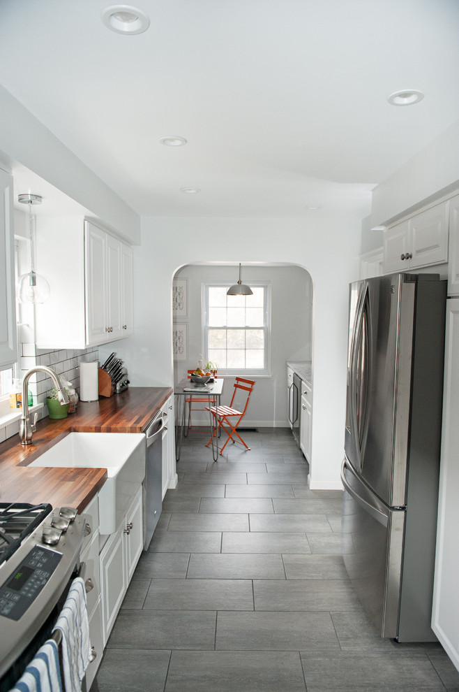 Mid-sized contemporary galley separate kitchen in Cincinnati with a farmhouse sink, raised-panel cabinets, white cabinets, wood benchtops, white splashback, ceramic splashback, stainless steel appliances, porcelain floors and no island.