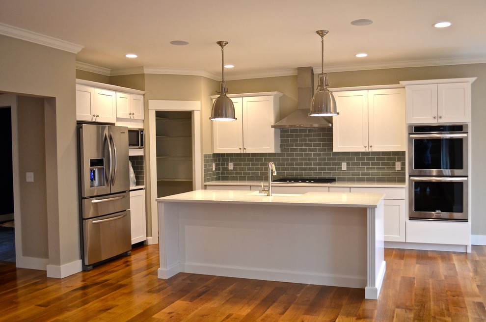Mid-sized transitional l-shaped open plan kitchen in Providence with a farmhouse sink, shaker cabinets, white cabinets, quartzite benchtops, grey splashback, subway tile splashback, stainless steel appliances, medium hardwood floors, with island and brown floor.