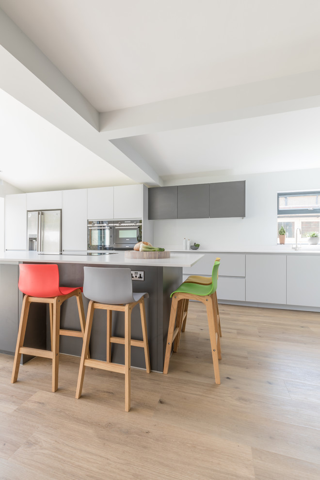 This is an example of a mid-sized contemporary galley open plan kitchen in Sussex with an integrated sink, flat-panel cabinets, grey cabinets, solid surface benchtops, stainless steel appliances, with island and white benchtop.