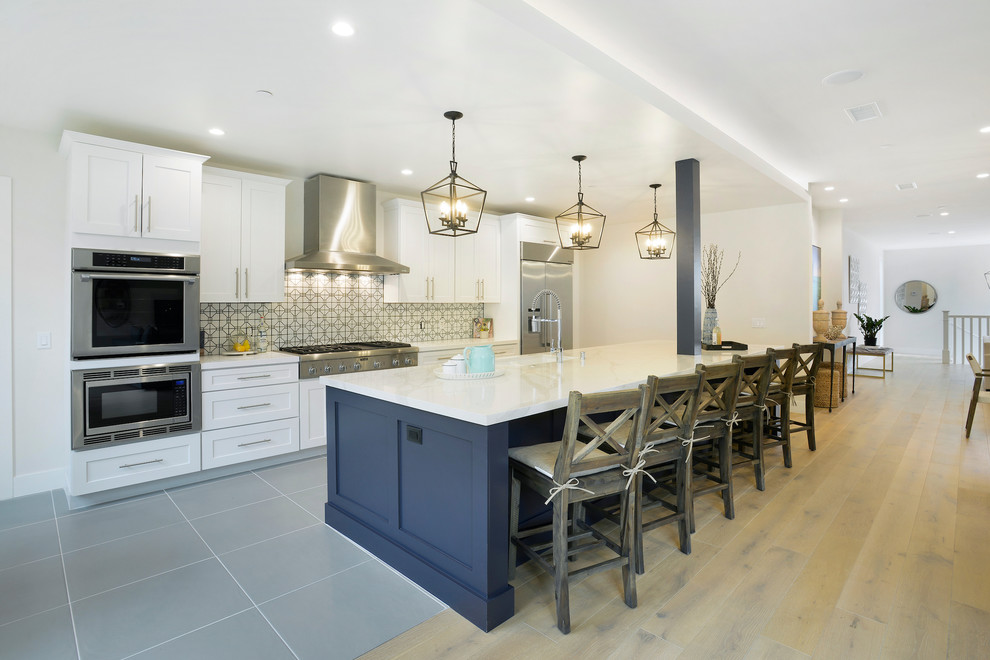 This is an example of a mid-sized beach style single-wall open plan kitchen in Orange County with an undermount sink, shaker cabinets, white cabinets, multi-coloured splashback, ceramic splashback, stainless steel appliances, porcelain floors, with island, grey floor and white benchtop.