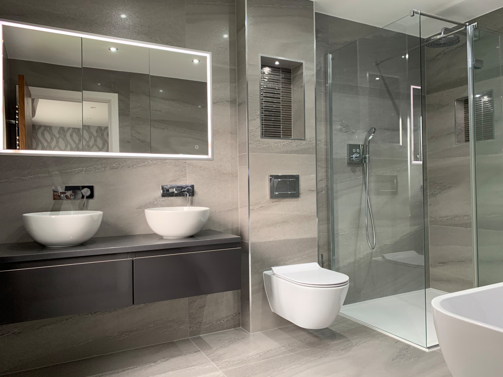 This is an example of a large contemporary ensuite bathroom in London with flat-panel cabinets, grey cabinets, a freestanding bath, a walk-in shower, a wall mounted toilet, grey tiles, porcelain tiles, grey walls, porcelain flooring, a vessel sink, quartz worktops, grey floors, an open shower, black worktops, an enclosed toilet, double sinks and a floating vanity unit.