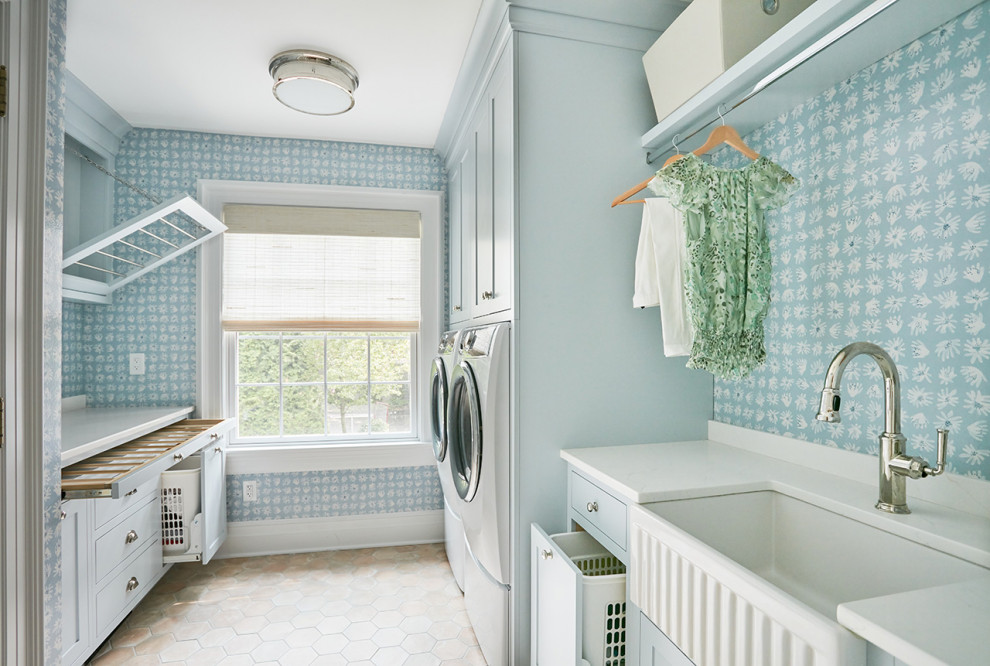 Design ideas for a mid-sized transitional galley laundry room in New York with a farmhouse sink, shaker cabinets, blue cabinets, solid surface benchtops, blue walls, porcelain floors, a side-by-side washer and dryer, white floor, white benchtop and wallpaper.