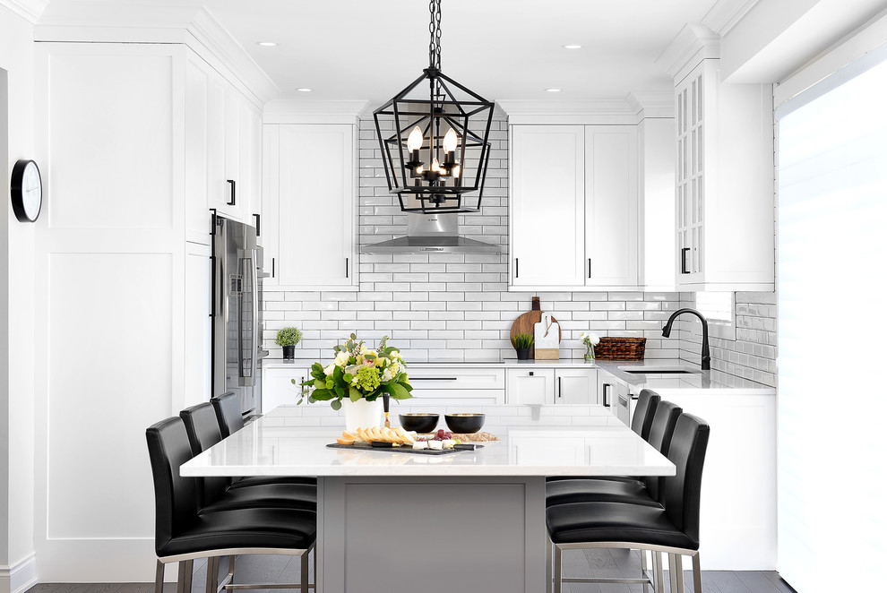 Photo of a transitional u-shaped eat-in kitchen in Toronto with an undermount sink, shaker cabinets, white cabinets, white splashback, subway tile splashback, stainless steel appliances, with island, black floor and white benchtop.