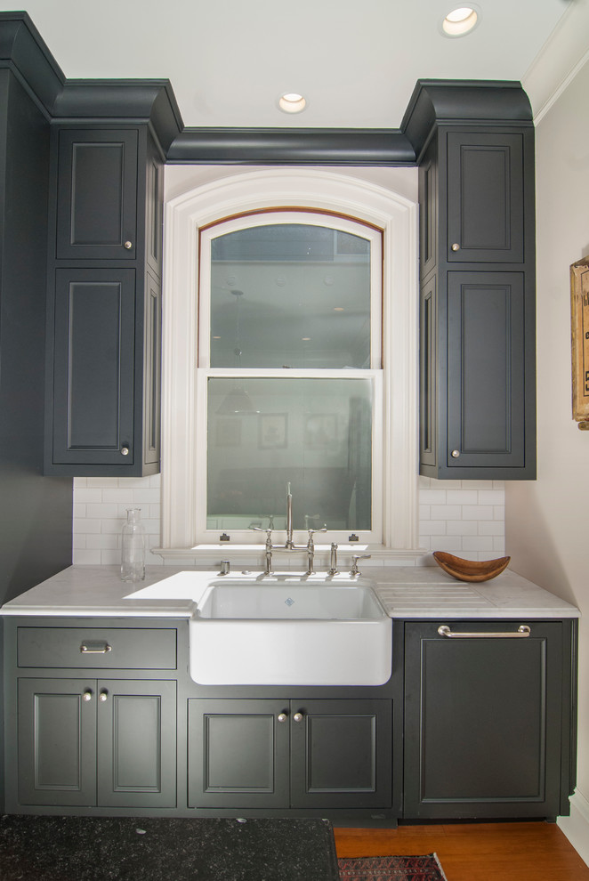 Photo of a mid-sized traditional separate kitchen in San Francisco with a farmhouse sink, flat-panel cabinets, black cabinets, marble benchtops, white splashback, subway tile splashback, panelled appliances, light hardwood floors and with island.