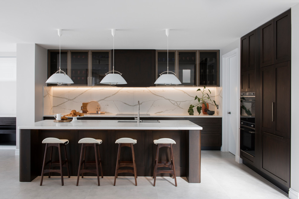 Mid-sized contemporary galley open plan kitchen in Sydney with an undermount sink, shaker cabinets, dark wood cabinets, marble benchtops, white splashback, stone slab splashback, black appliances, porcelain floors, with island and grey floor.