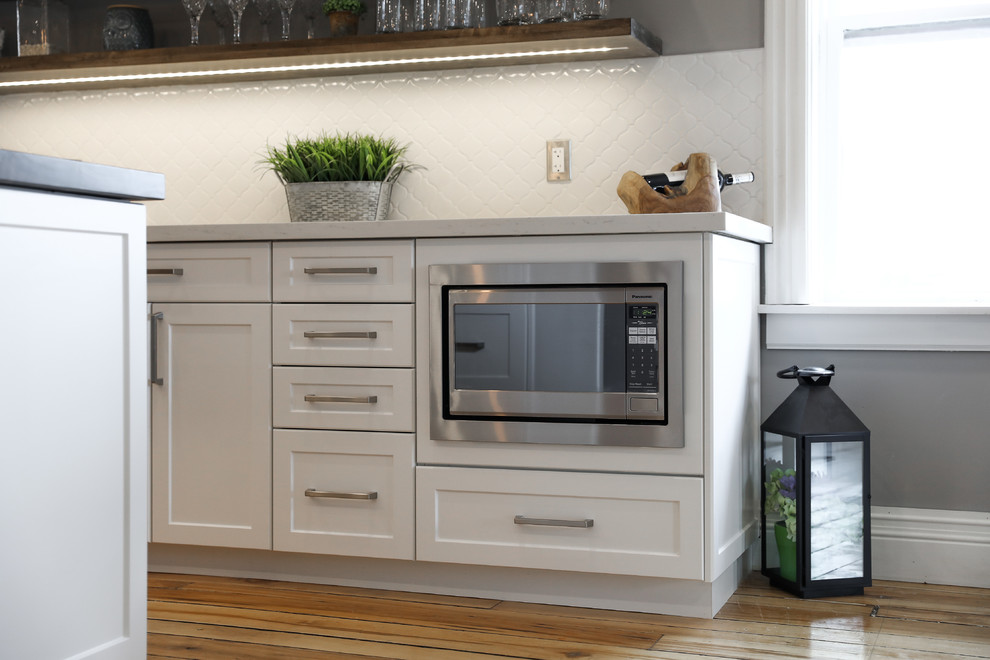 Example of a mid-sized transitional single-wall medium tone wood floor and brown floor eat-in kitchen design with a farmhouse sink, shaker cabinets, white cabinets, white backsplash, stainless steel appliances, an island and white countertops