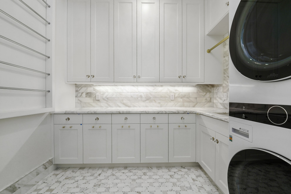 Inredning av en stor grå l-formad grått tvättstuga enbart för tvätt, med en undermonterad diskho, luckor med infälld panel, vita skåp, marmorbänkskiva, grått stänkskydd, stänkskydd i marmor, vita väggar, marmorgolv, en tvättpelare och grått golv