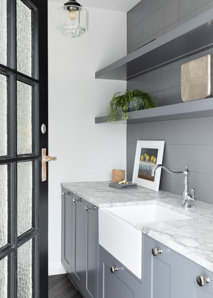 Photo of a mid-sized country single-wall dedicated laundry room in Auckland with a farmhouse sink, shaker cabinets, grey cabinets, quartzite benchtops, grey splashback, ceramic splashback, white walls, dark hardwood floors, a concealed washer and dryer, black floor and white benchtop.