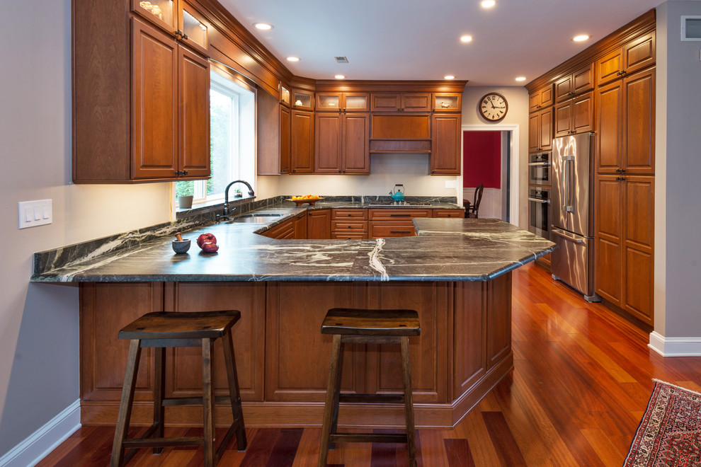 This is an example of a large traditional u-shaped eat-in kitchen in Philadelphia with an undermount sink, raised-panel cabinets, medium wood cabinets, soapstone benchtops, black splashback, stone slab splashback, stainless steel appliances, medium hardwood floors and a peninsula.