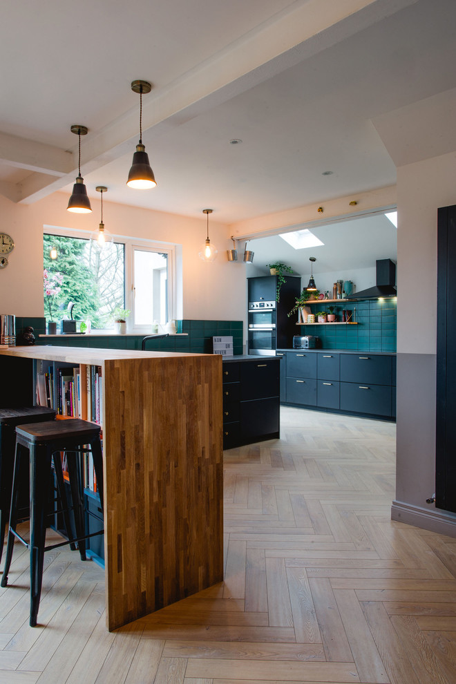 Contemporary l-shaped eat-in kitchen in Other with flat-panel cabinets, black cabinets, wood benchtops, green splashback, subway tile splashback, stainless steel appliances, light hardwood floors, a peninsula, beige floor and brown benchtop.